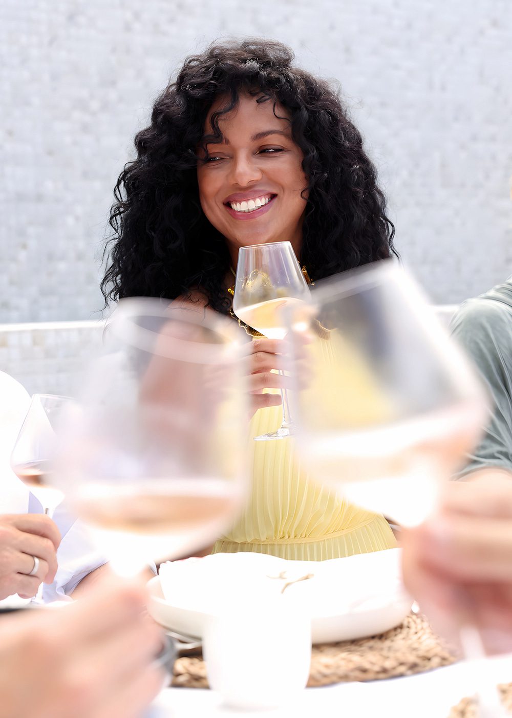 A smiling woman enjoys a glass of Hanikon Rosé wine at an outdoor gathering with friends. Her cheerful expression, surrounded by blurred wine glasses in the foreground, captures the essence of Hanikon's social and celebratory wine experience.