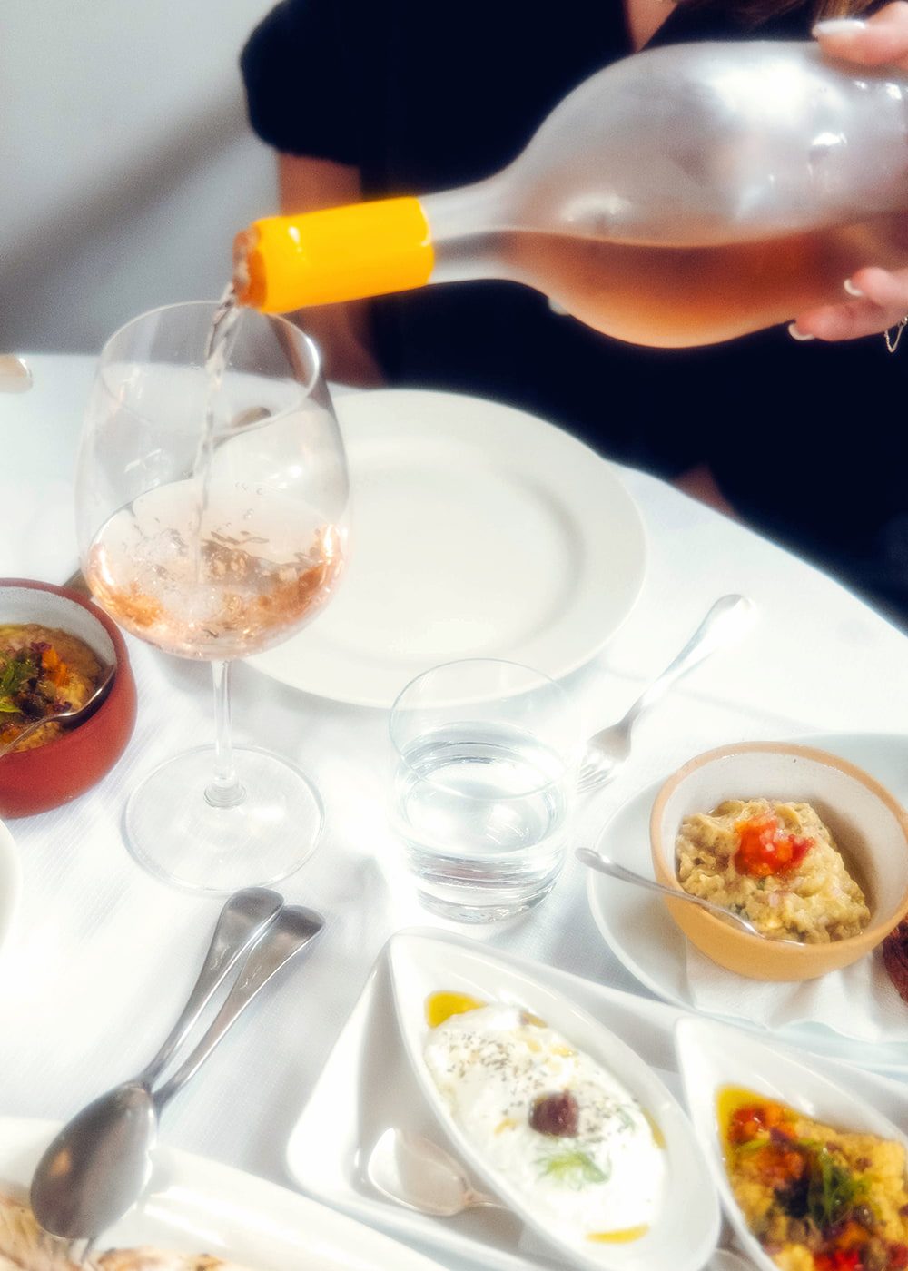 A bottle of Hanikon Côtes de Provence Rosé being poured into a wine glass at a restaurant table, surrounded by an array of Mediterranean dishes. The scene captures the essence of dining with Hanikon Rosé, enhancing the meal with its fresh and vibrant flavor.