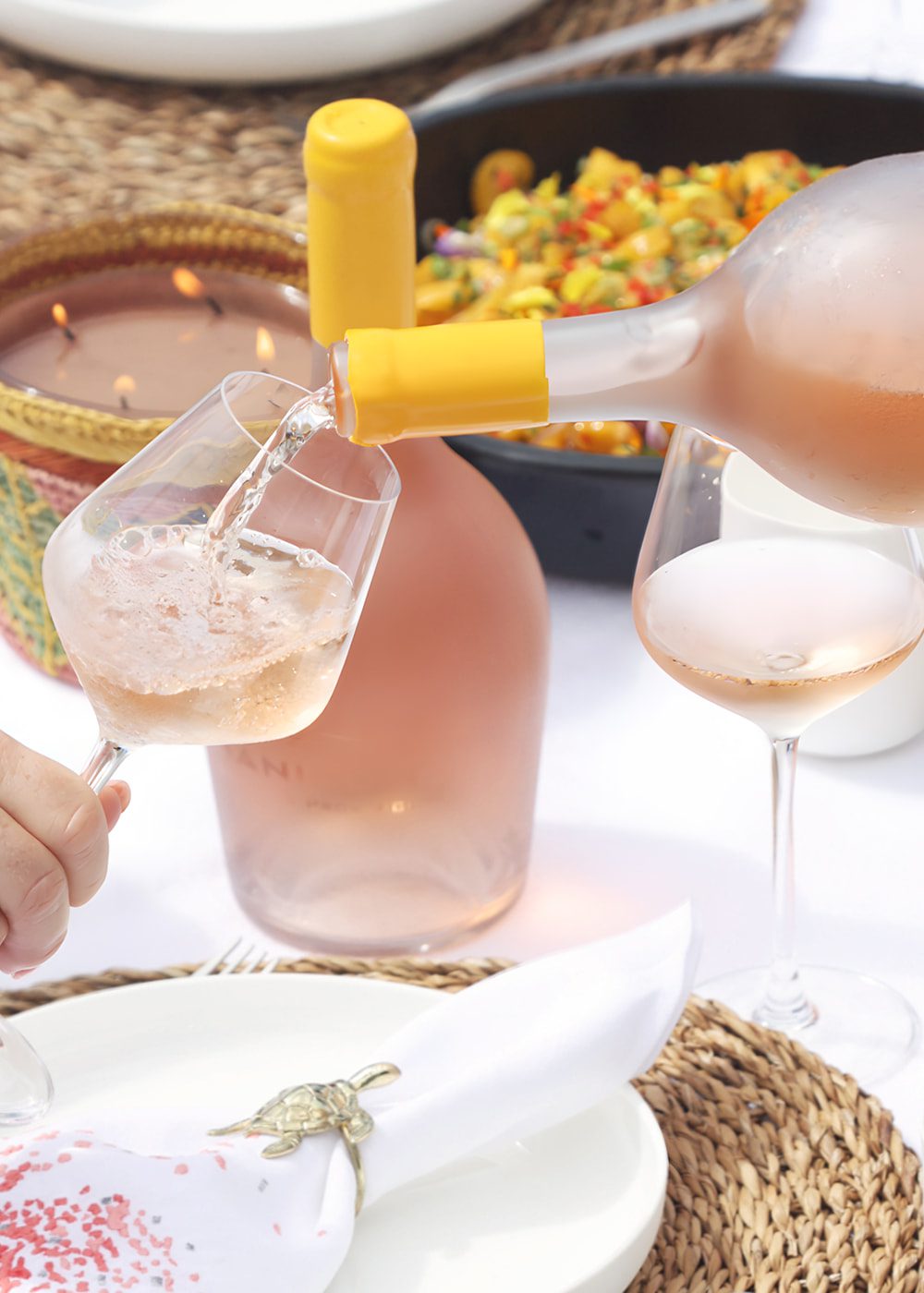 A glass of Hanikon Rosé wine being poured at a beautifully set table, featuring a large bottle of rosé, a woven placemat, and colorful food in the background. The relaxed, elegant dining setup highlights the perfect pairing of Hanikon Rosé with a summer meal and gatherings.