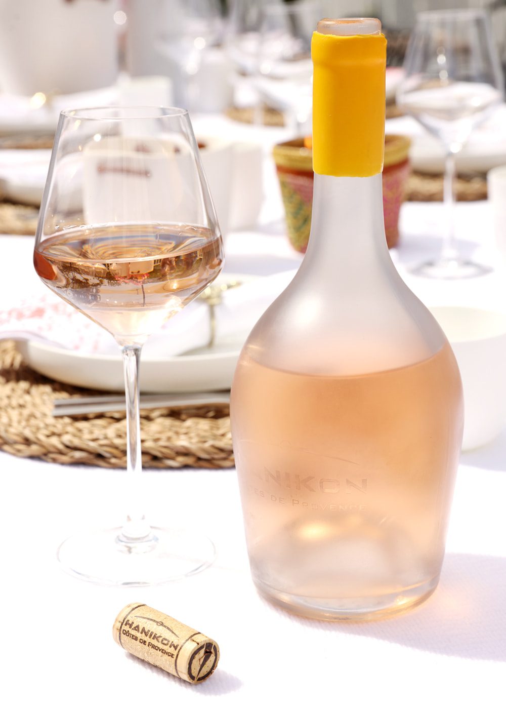 A bottle of Hanikon Côtes de Provence Rosé sits on a beautifully set table, accompanied by a filled wine glass and a cork. The outdoor dining setup reflects the refreshing, celebratory nature of Hanikon Rosé, ideal for enjoying with friends and family.